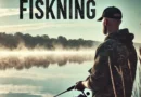 A person in camouflage attire fishing by a misty lake at dawn, with the word "FISKNING" written at the top. The scene is calm and serene, with soft light and reflections on the water, evoking a peaceful outdoor experience.