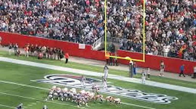 An American football team prepares for a field goal attempt during a game, with players lined up near the end zone. The goalposts and a large, excited crowd in the stadium are visible in the background.