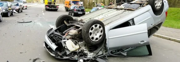 A flipped silver car lying upside down on a road after a severe accident. Debris is scattered around the vehicle, and other cars, along with emergency vehicles, are visible in the background. The scene shows the aftermath of a serious traffic collision.