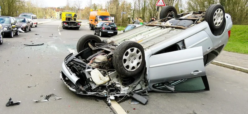 A flipped silver car lying upside down on a road after a severe accident. Debris is scattered around the vehicle, and other cars, along with emergency vehicles, are visible in the background. The scene shows the aftermath of a serious traffic collision.