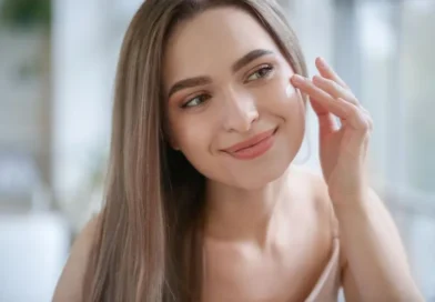 A close-up of a woman with glowing, healthy skin applying moisturizer to her cheek. She smiles gently, appearing relaxed, in a well-lit room with a soft, blurred background.