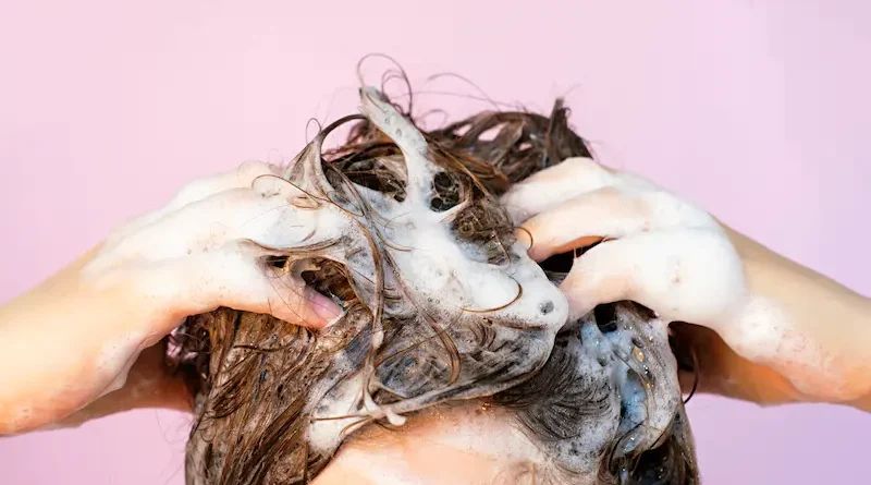 A person washing their hair with shampoo, creating a thick lather on their scalp. Their hands are visible massaging the suds into the hair. The background is a soft lilac color.