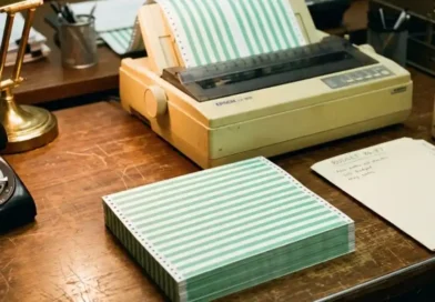 A stack of green and white striped continuous-feed printer paper next to a vintage dot-matrix printer in a 1980s office setting