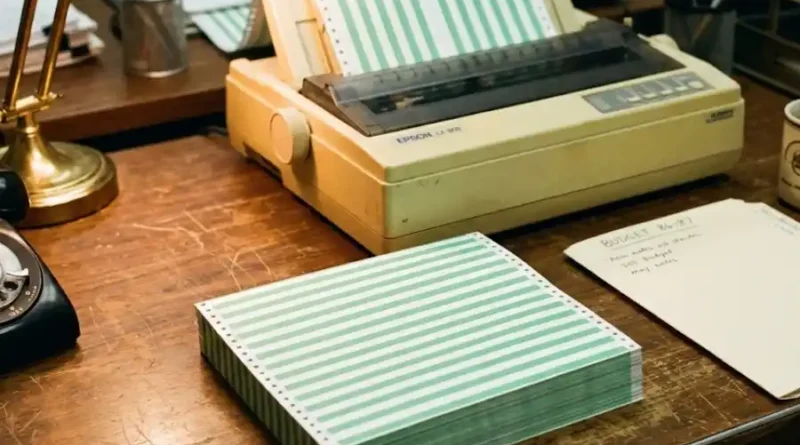 A stack of green and white striped continuous-feed printer paper next to a vintage dot-matrix printer in a 1980s office setting